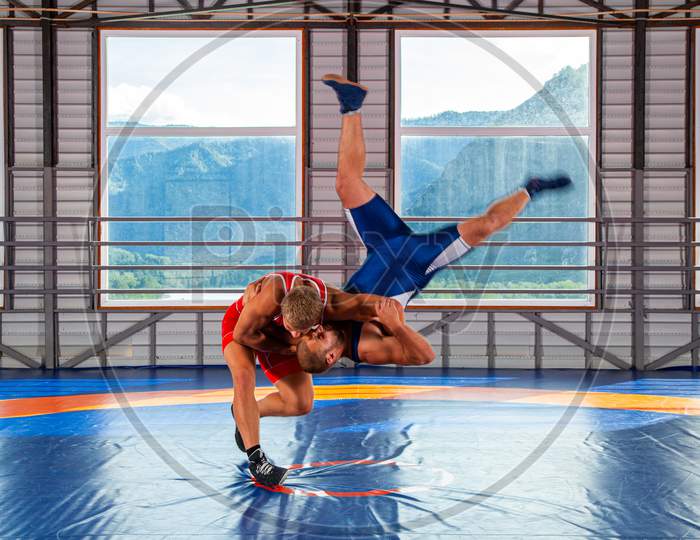 Greco-Roman Wrestling Training, Grappling. Two Greco-Roman  Wrestlers In Red And Blue Uniform Making A Thigh Throw  On A Wrestling Carpet In The Gym.The Concept Of Male Wrestling And Resistance