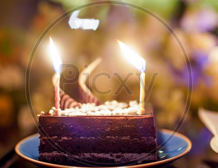 Shot Of Cake With Sparkling Candles With Flames And Sparks Flying Out With An Out Of Focus Background Of Flowers, Lights Showing A Birthday Celebration, Wedding Or An Anniversary