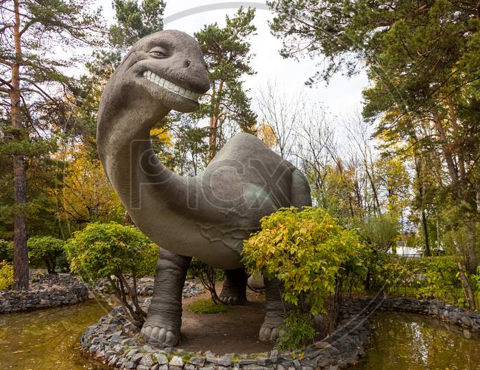 A Large Gray Adult Brachiosaurus Looks Towards The Frame And Stands On The Stone Bank Of The River, Next To Small Trees And Shrubs On A Warm Autumn Day. Dinosaur World