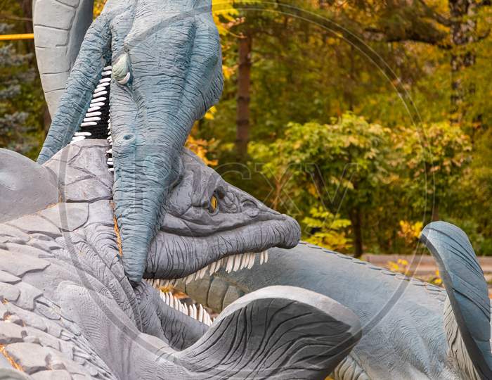 A Large Gray Adult Elasmosaurus Attacks An Ancient Crocodile On A Stone Bank Of A River, Next To Small Trees And Shrubs On A Warm Autumn Day. Dinosaur World