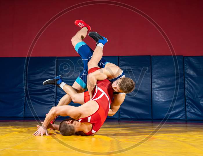 Two Strong Wrestlers In Blue And Red Wrestling Tights Are Wrestlng  On A Wrestling Carpet In The Gym. Young Man Doing Grapple.