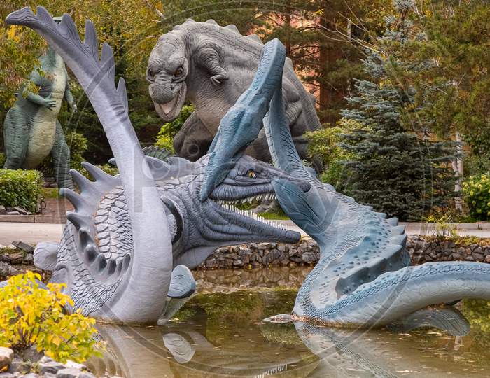 A Large Gray Adult Elasmosaurus Attacks An Ancient Crocodile On A Stone Bank Of A River, Next To Small Trees And Shrubs On A Warm Autumn Day, In The Background A Tyronasaurus. Dinosaur World
