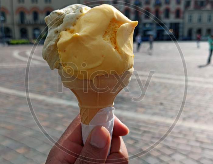 Close Up Of Hand Holding A Delicious Looking Saffron And Coffee Ice Cream Cone In Front Of European Building Exterior Located In The Center Of Krakow, Poland, Europe. Travel Concept During Summer