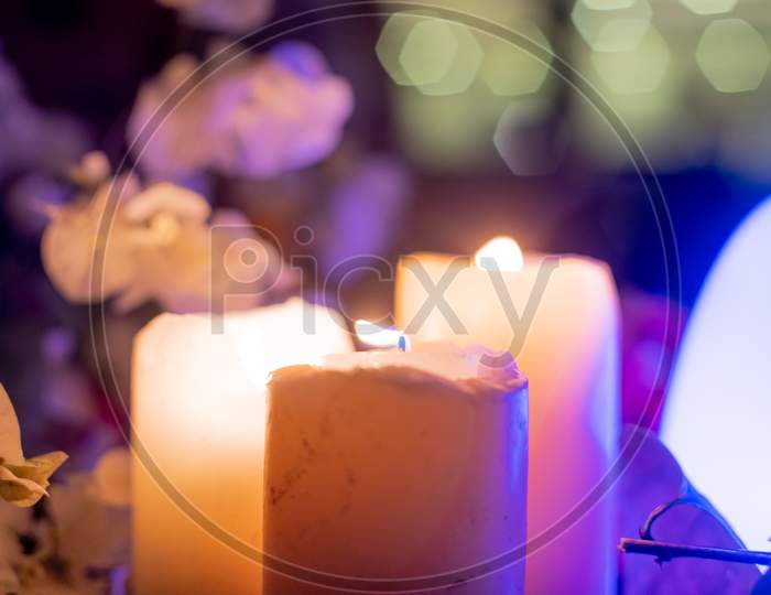 Zoomed In Shot Of Large Thick Wax Candles With Flames Lit And Colorful Flowers All Round And Led Lights Flickering Showing The Table Decorations At A Valentine , Wedding , Engagement Dinner At Night With An Intimate Beautiful Display