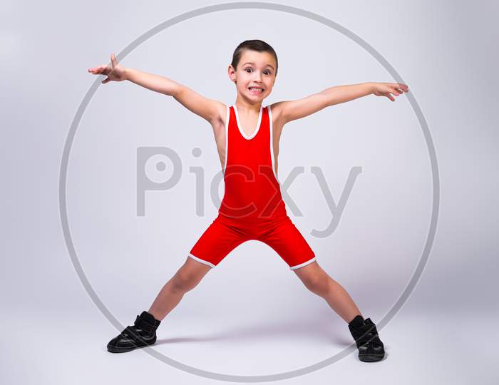 Little Funny Boy In A Red Sports Ortsovskoy Leotard And Wrestling Boots Holds Arms And Legs In Different Directions And Smiles At His Achievements On A White Isolated Background.  The Concept Of A Little Fighter Athlete.