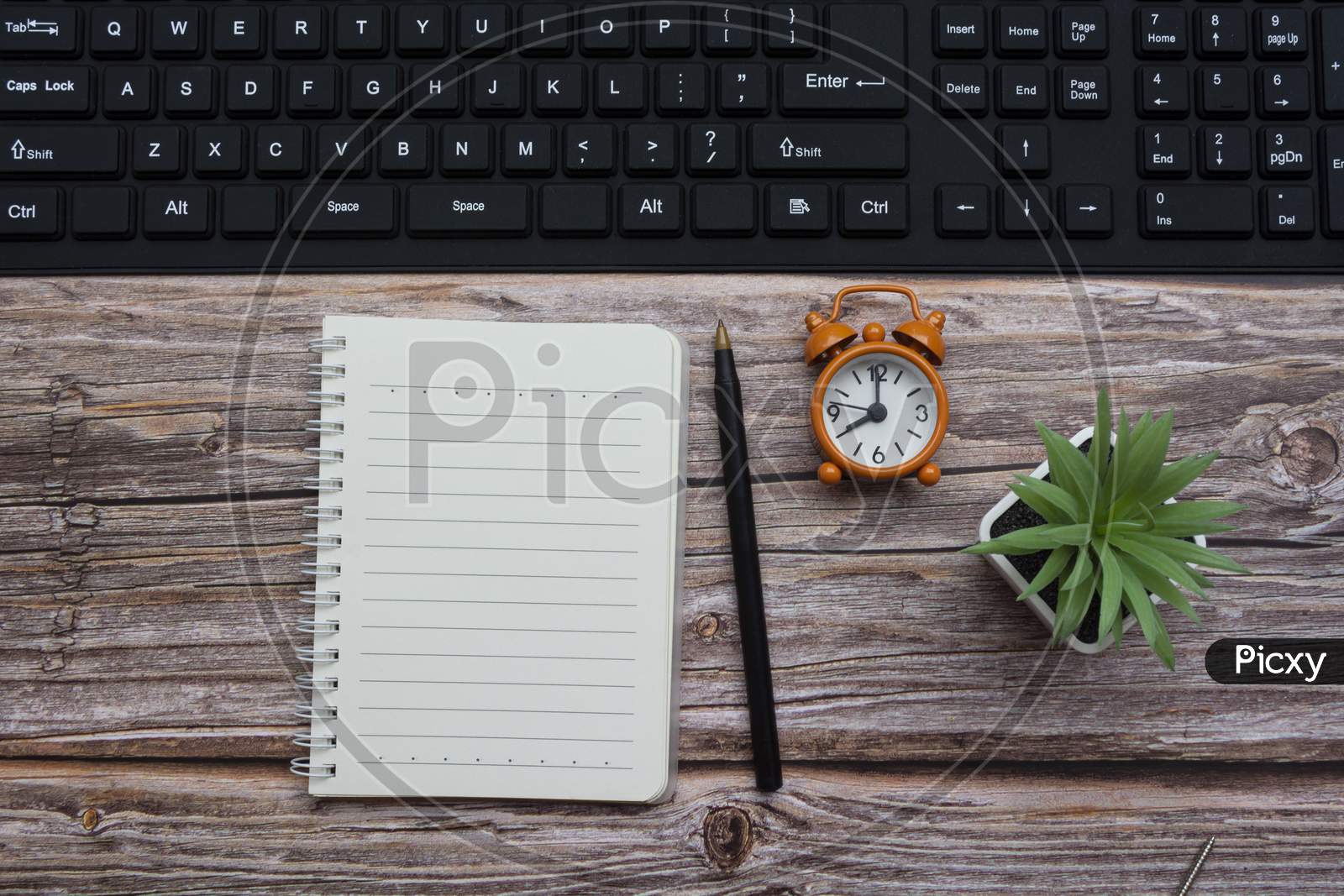 Notepad With Pen, Alarm Clock, Potted Plan And Keyboard On Wooden Desk