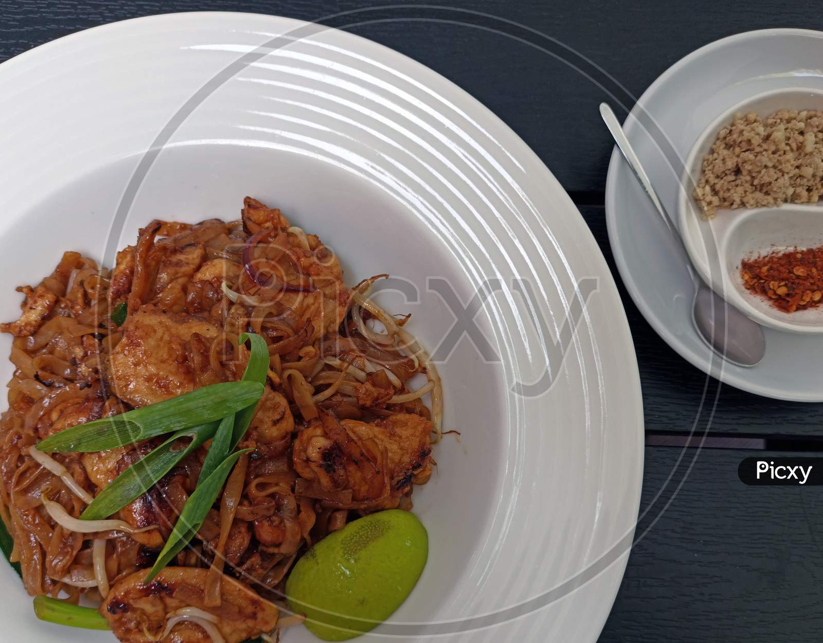 Pad Thai Noodles With Fried Chicken, Mixed Green Leaves, Chopped Peanuts, Shallots & Lime Wedge On White Plate Against Wooden Background Served Hot With Spices And Chillies