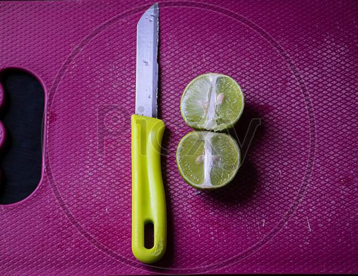 Stock Photo Of Fresh Juicy Cut Lemon Kept On Purple Color Chopping Board With Yellow Color Knife, Focus On Object. Blur Background.