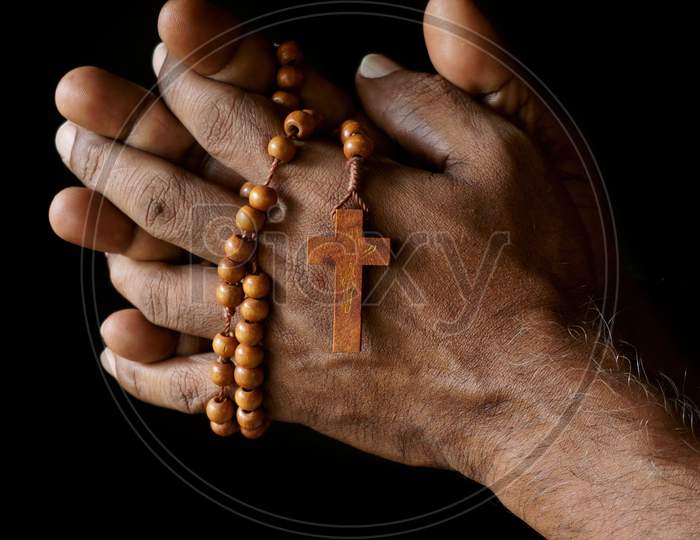 Praying Hand Of An Old Indian Catholic Man With Wooden Rosary Isolated On A Plain Black Background.