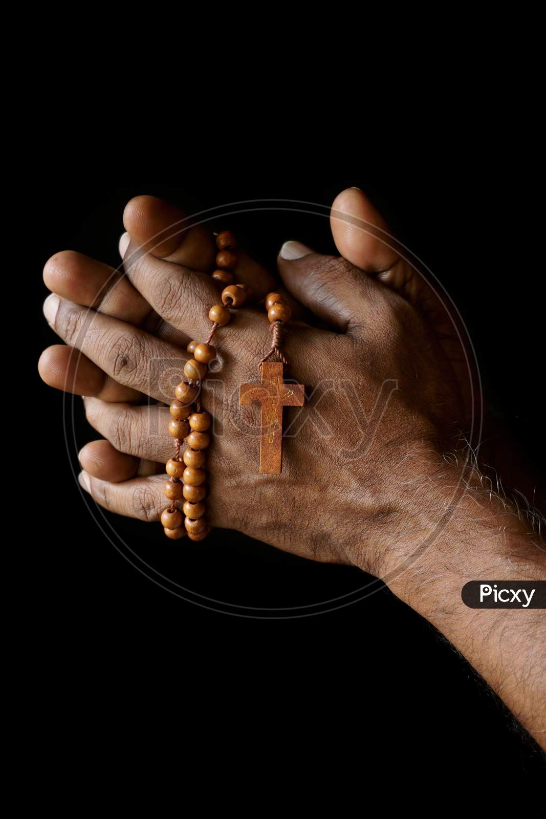 Praying Hand Of An Old Indian Catholic Man With Wooden Rosary Isolated On A Plain Black Background.