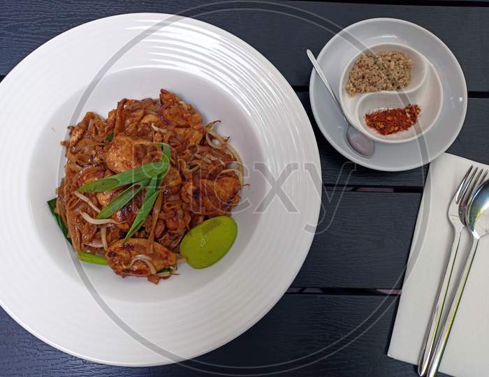 Pad Thai , Stir Fried Dish Made With Noodles, Chicken, Vegetables, Peanuts And Slice Of Lemon On A Plate Against Wooden Pattern Background And Fork Utensils.