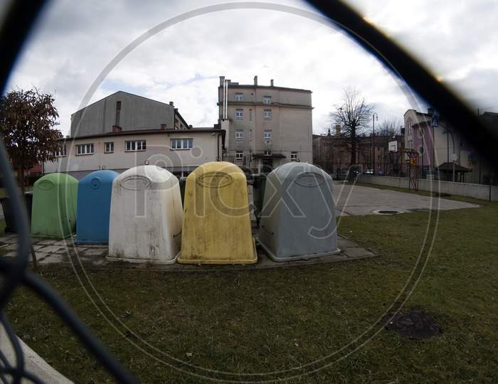 Wide Angle View Through The Fence Of Bunch Of Trash Containers Or Dust Bins For Garbage Placed In The School Premises In Southern Poland Town. Europe