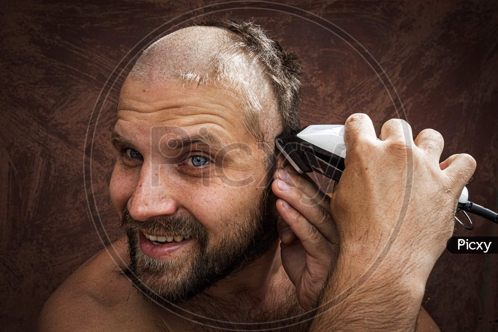 Caucasian Man Trying To Shave His Head With An Electric Razor. A Brutal Bald Man Holds A Razor In His Hand And Shaves Stubble On A Metal Background