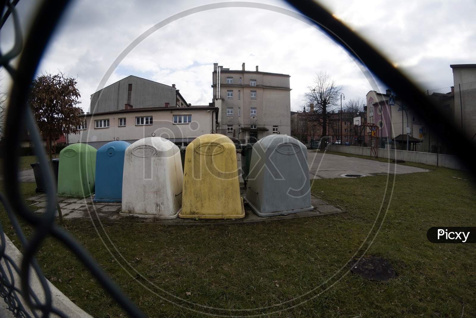 Wide Angle View Through The Fence Of Bunch Of Trash Containers Or Dust Bins For Garbage Placed In The School Premises In Southern Poland Town. Europe