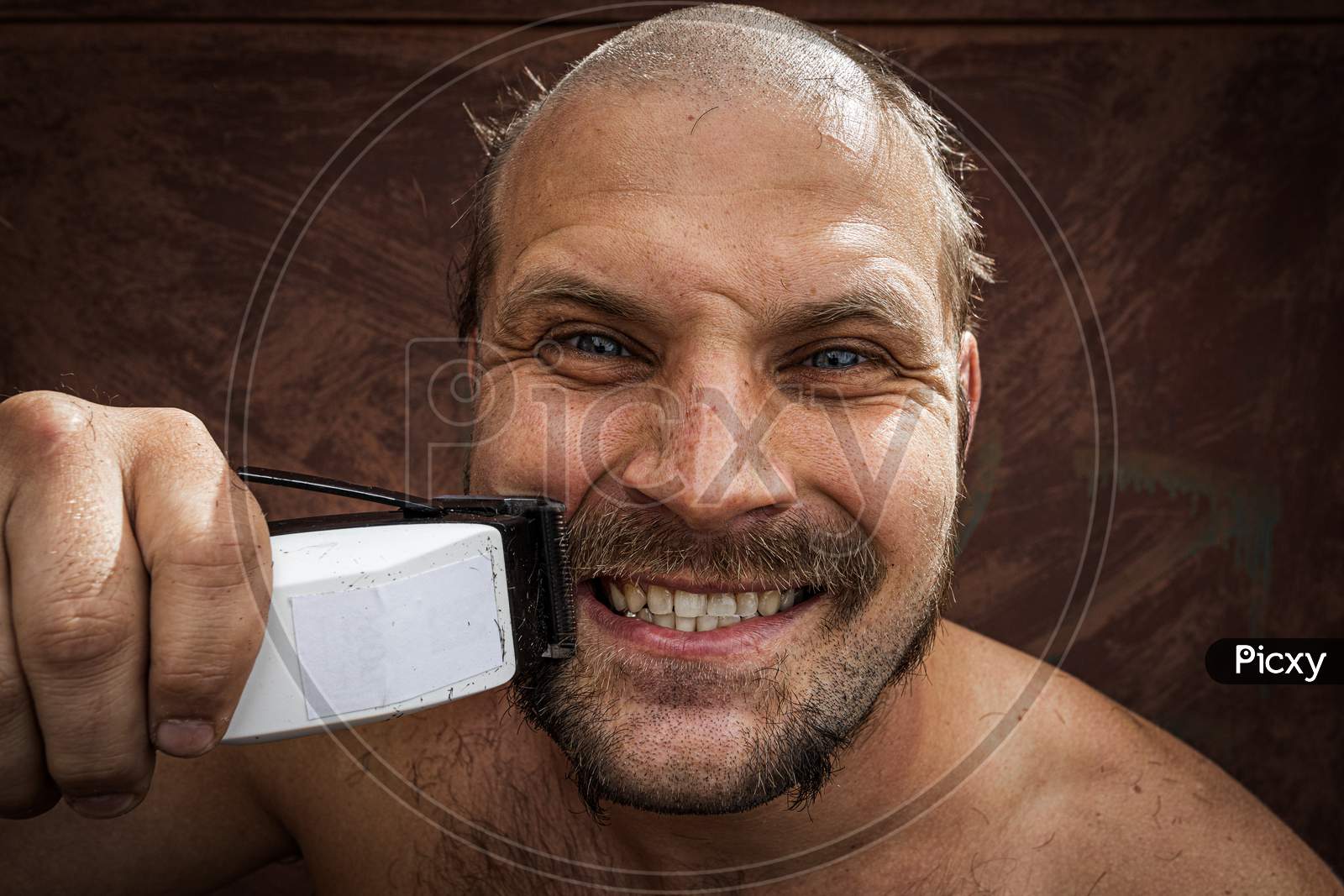 Adult Cheerful Man With A Beard Shaves His Beard At Home. The Guy Looks In The Bathroom Mirror And Uses The Electric Trimmer. Front View