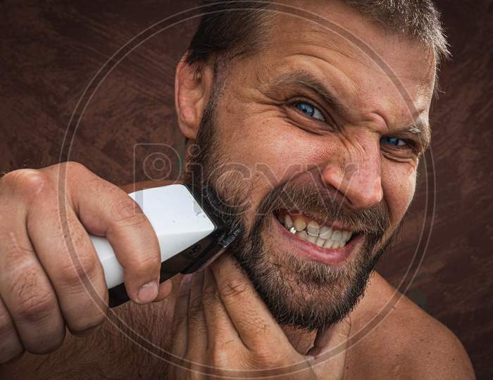 Close-Up Portrait Of Handsome Shirtless Man Shaving His Beard With An Electric Razor And Gritting His Teeth, Against Brutal Background. Concept Of Male Home Care Without Beauty Salons