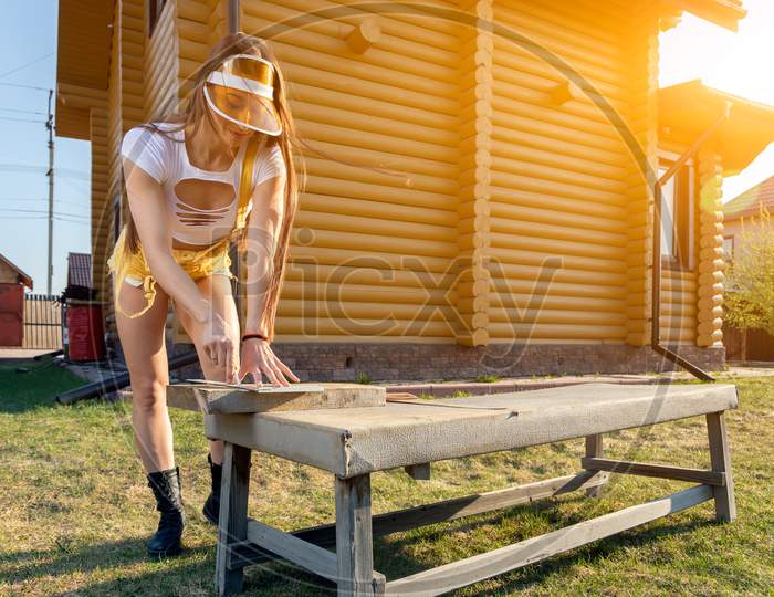 Young Athletic Woman In Jeans And Crop Top Is Sawing Wood In A Rustic Atmosphere On A Warm Summer Day, In The Background A Wooden House And A Lawn