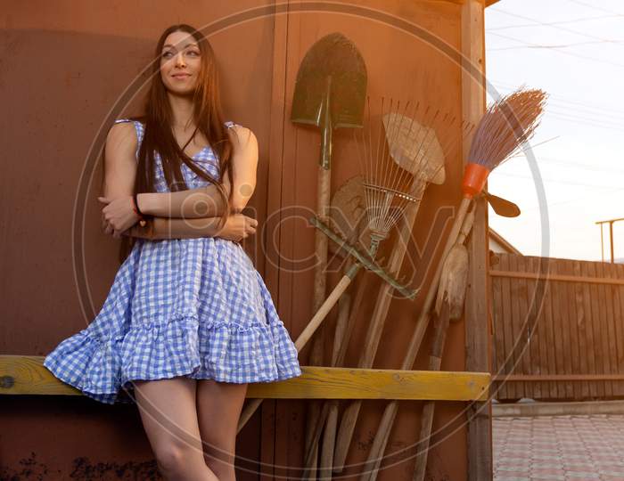 A Pretty Fresh Dark-Haired Young Woman In A Summer Short Demonstrates Her Athletic Body Against The Background Of A Fence Next To Summer Tools, Shovels, Rakes. The Concept Of Summer Vacations In The Village. Fitness Model Posing For Summer
