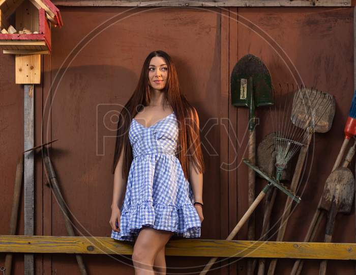 A Pretty Fresh Dark-Haired Young Woman In A Summer Short Demonstrates Her Athletic Body Against The Background Of A Fence Next To Summer Tools, Shovels, Rakes. The Concept Of Summer Vacations In The Village. Fitness Model Posing For Summer