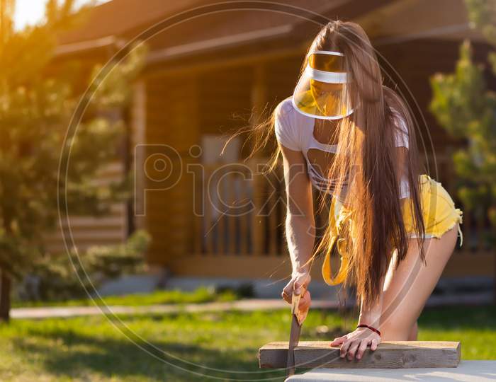 A Pretty Woman Carpenter In Stylish Lazy Clothes And A Visor, Cuts A Wooden Board With A Saw. A Young Woman In Short Shorts, A Top, Is Engaged In Farm Chores, Works With A Tree. Strong Woman Lumberjack