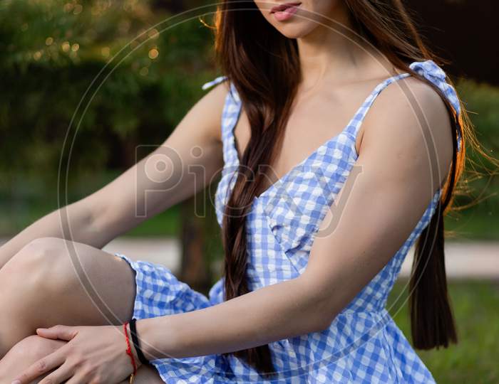 A Young Woman  Sits Relaxed In A Checkered Short Dress   On The Green Grass In The Park On A Warm Day. The Concept Of Summer Vacation In The Village And Live Style