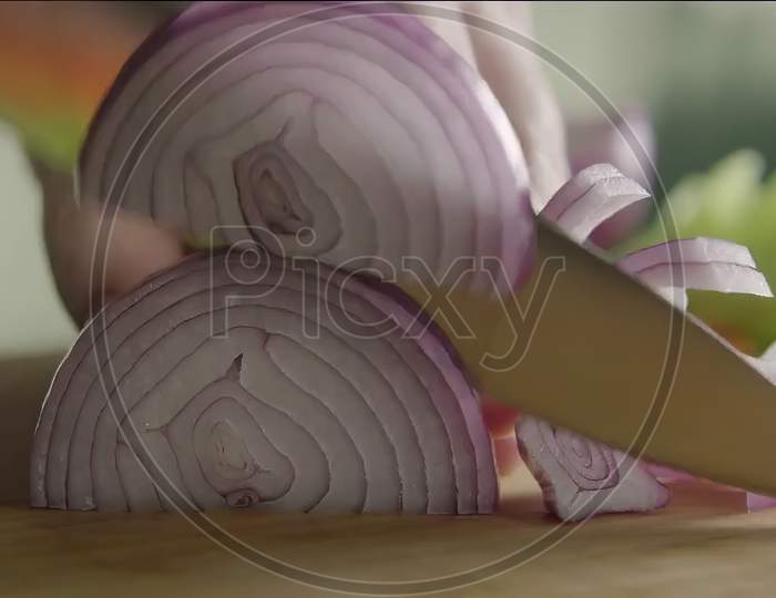 Closeup of hand chopping onion in to thin slices with a sharp knife on a wooden base.