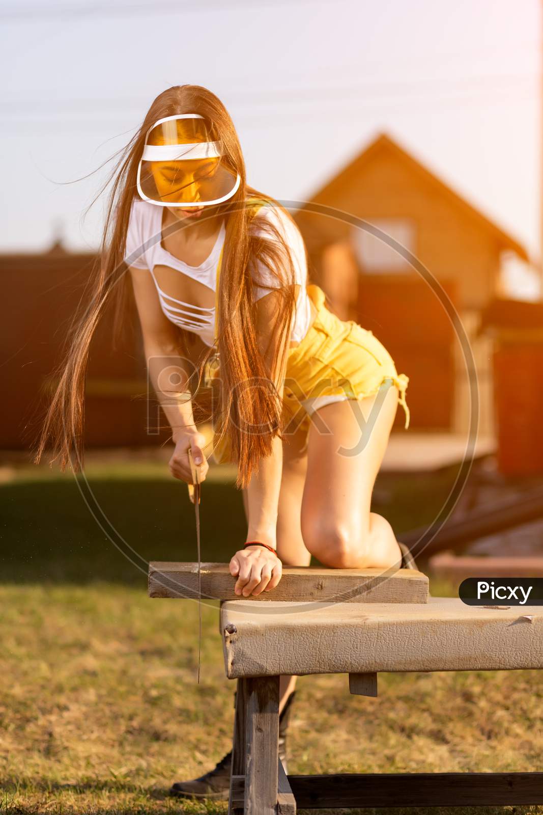 Young Athletic Woman In Jeans And Crop Top Is Sawing Wood In A Rustic Atmosphere On A Warm Summer Day, In The Background A Wooden House And A Lawn