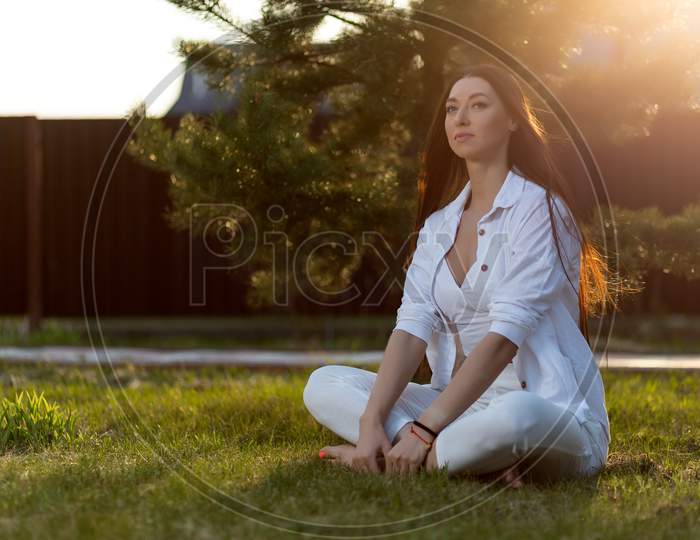 A Young Woman  Sits Relaxed In A White Top, Shirt And Jeans On The Green Grass In The Park On A Warm Day. The Concept Of Summer Vacation In The Village And Live Style