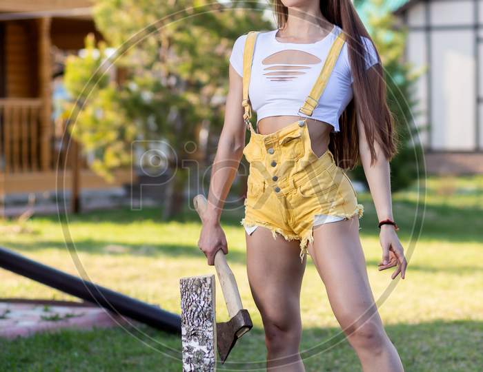 Fashionable Portrait Of Young Woman, Short Shorts And Top, Posing, Going To Chop Wood With An Ax In A Village With A Lawn On A Summer Day. Portrait Of A Joyful Woman