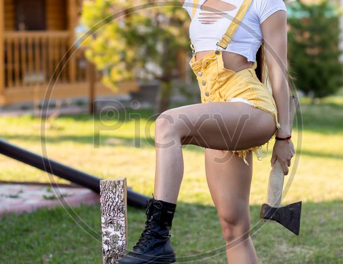 Young Athletic Woman In Jeans And Crop Top Holds An Ax In Her Hands And Is About To Chop Wood In A Rustic Atmosphere On A Warm Summer Day. Fitness Woman With Ax