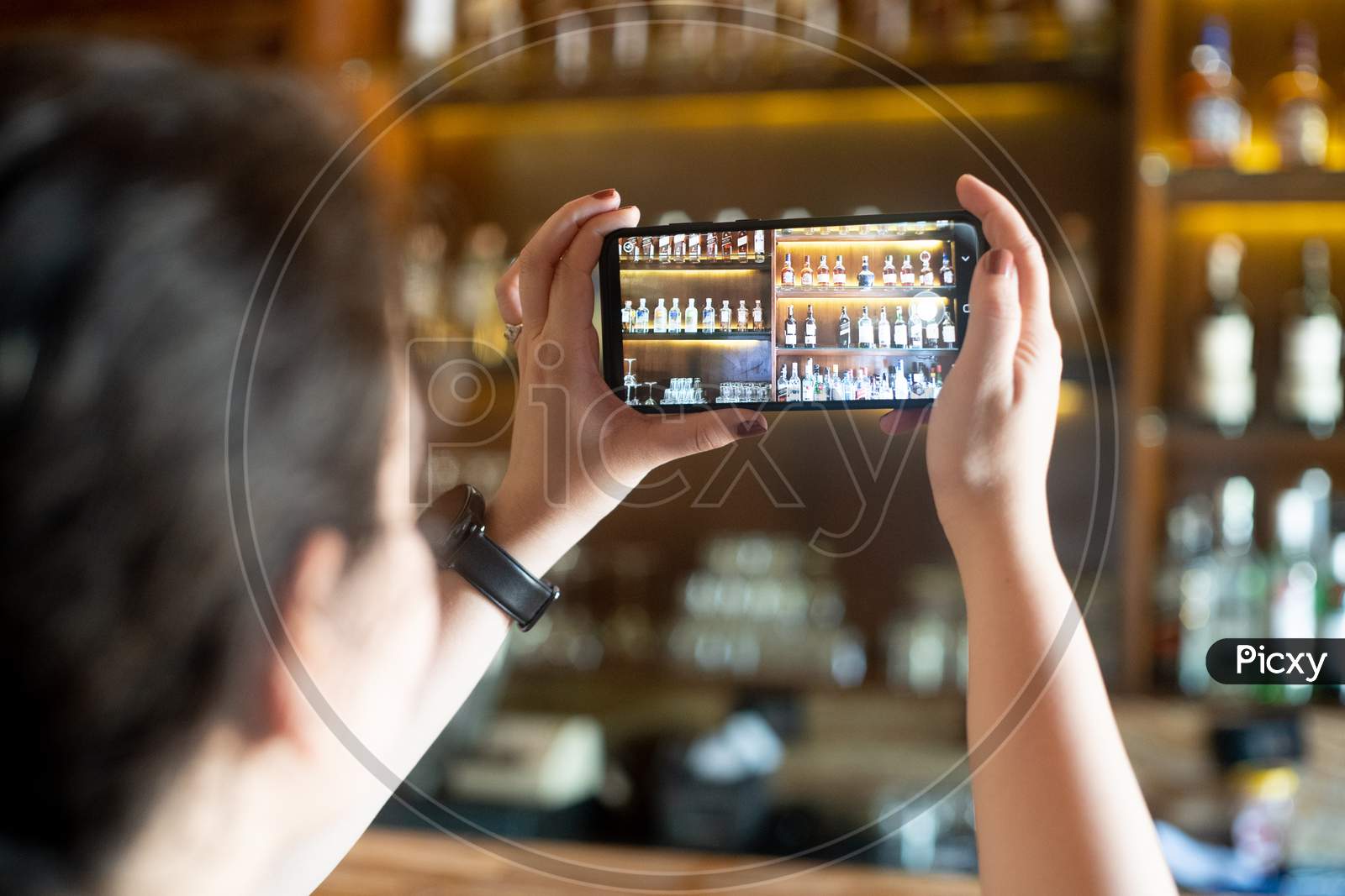 Young Indian Woman Taking A Picture Of A Bar Pub Shelf With Alcohol Bottles Stacked On Wooden Shelf With Phone In Landscape