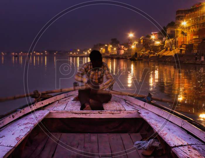 Varanasi, India - November 01, 2016: A Man Rowing A Wooden Paddle On A Boat Before Sunrise In Ganges River Against Illuminated Varanasi Holy City Located In The State Of Uttar Pradesh.