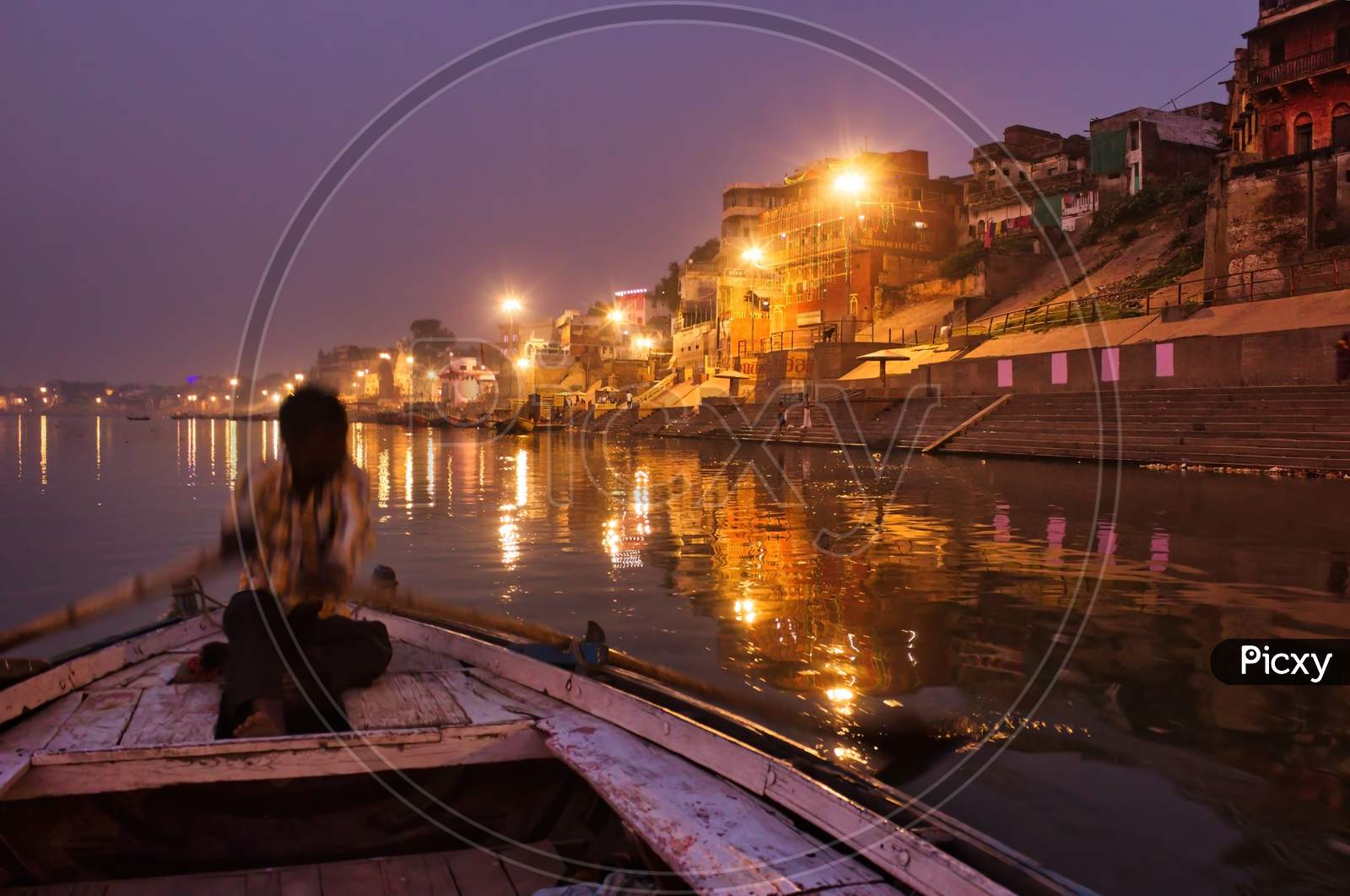 Varanasi, India - November 01, 2016: A Man Rowing A Wooden Paddle On A Boat Before Sunrise In Ganges River Against Illuminated Varanasi Holy City Located In The State Of Uttar Pradesh.