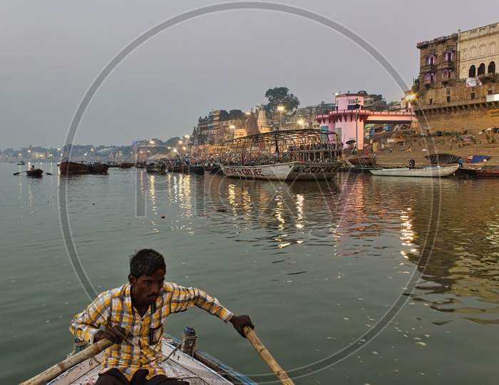 Varanasi, India - November 01, 2016: A Man Rowing A Wooden Boat Over River Ganga With Bamboo Paddles Against Famous Ghat In A State Of Uttar Pradesh.
