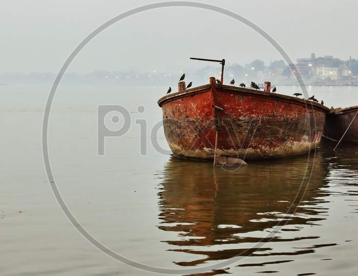 Bunch Of Birds Sitting On A Rusty Old Boat Against Mist Foggy Horizon In Varanasi Located In A State Of Uttar Pradesh, India