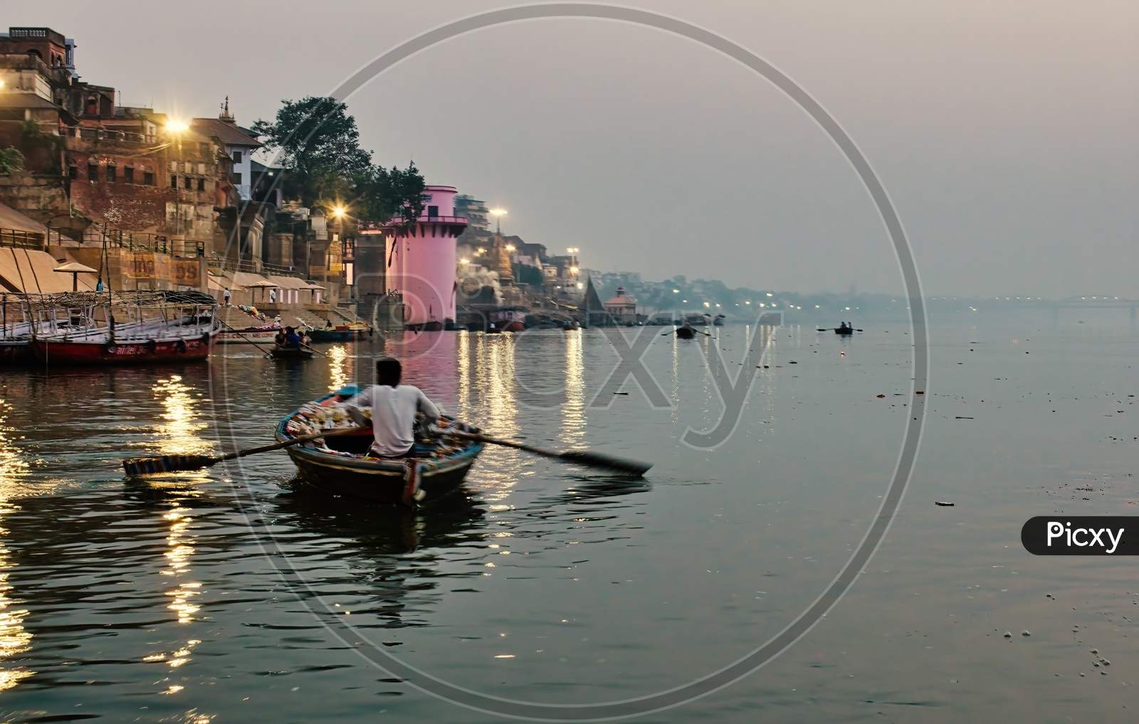 Varanasi, India - November 01, 2016: Wide Angle Of A Man Rowing A Wooden Boat Over River Ganga With Bamboo Paddles Against Famous Ghat And Old Architecture In A State Of Uttar Pradesh.