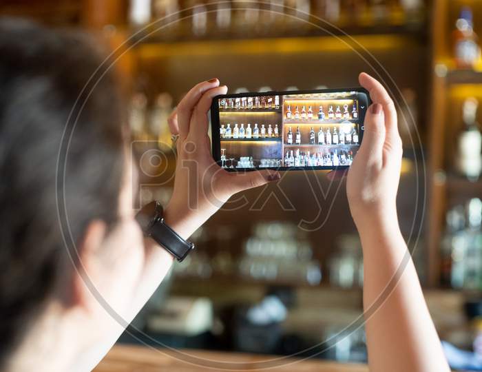 Young Indian Woman Taking A Picture Of A Bar Pub Shelf With Alcohol Bottles Stacked On Wooden Shelf With Phone In Landscape