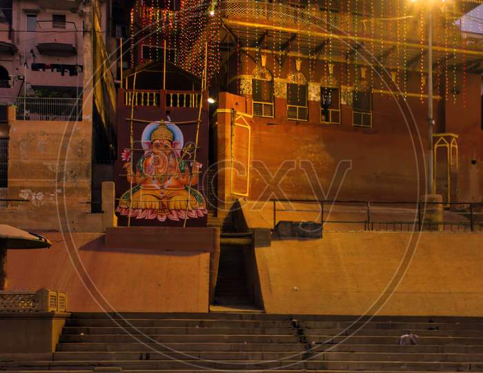 Varanasi, India - November 01, 2016: Bunch Of Wooden Boat Docked In Ganges River Next To Empty Ghat And Ancient Building Before Sunrise In State Of Uttar Pradesh. Hindu Holy Place With Ganesh Image