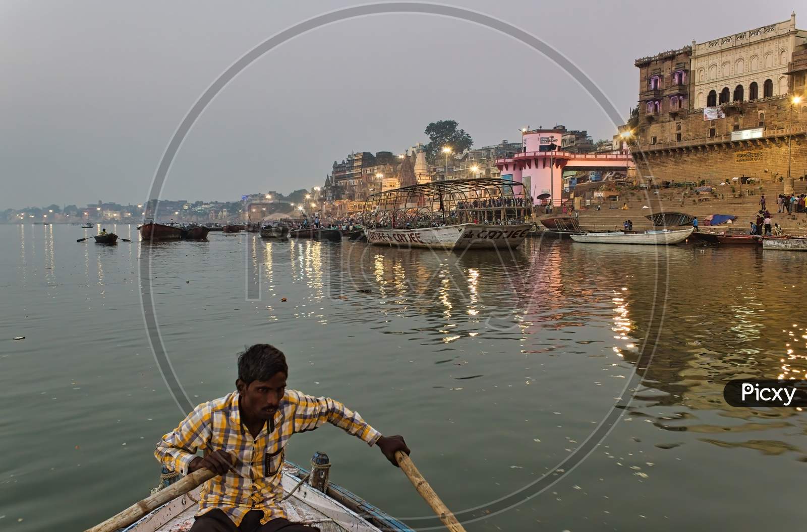 Varanasi, India - November 01, 2016: A Man Rowing A Wooden Boat Over River Ganga With Bamboo Paddles Against Famous Ghat In A State Of Uttar Pradesh.