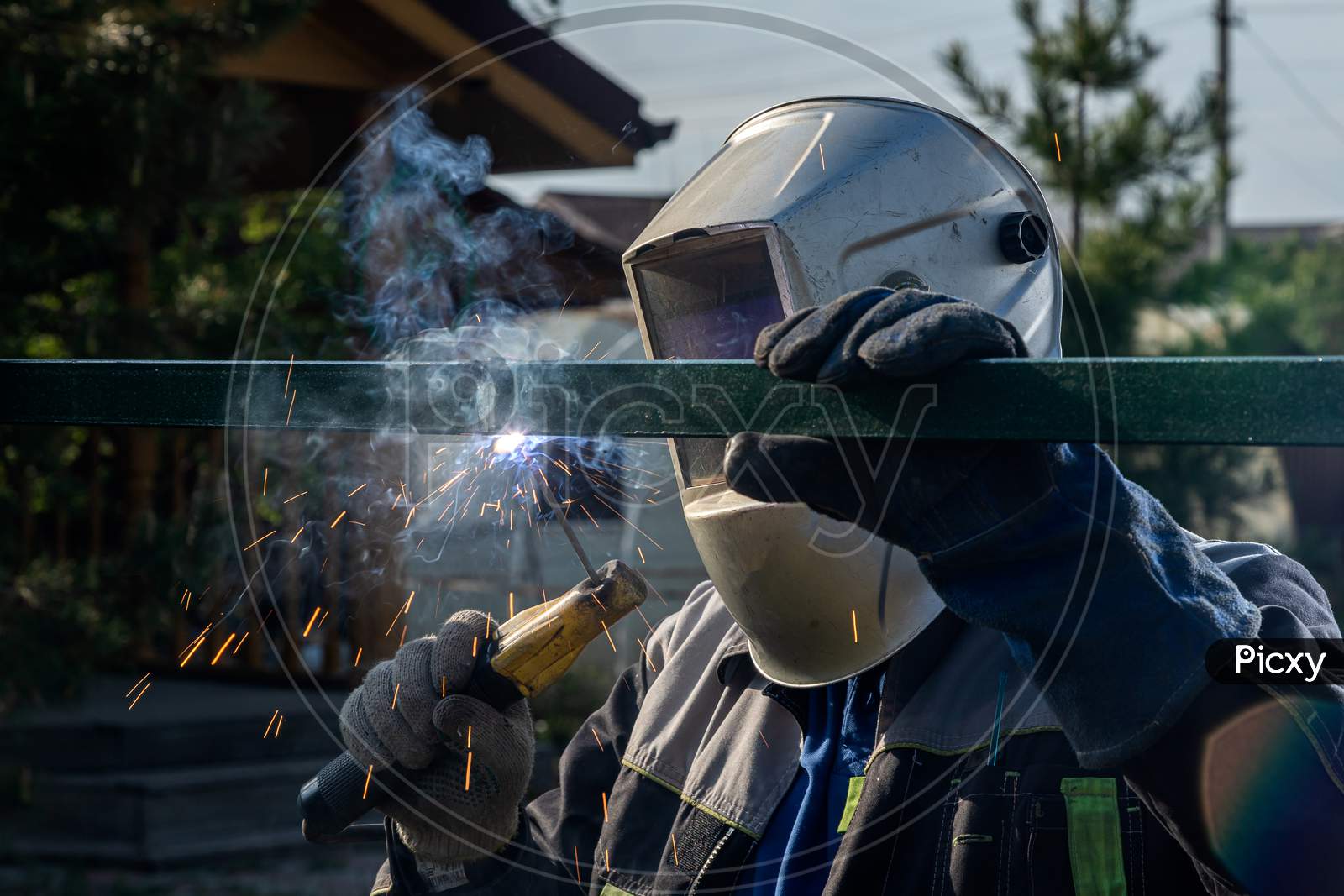 A Strong Man Is A Welder In A Welding Mask And Welders Leathers, A Metal Product Is Welded With A Welding Machine In The Outside,  Blue Sparks Fly To The Sides