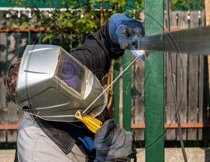 Close Up Of A Young  Man Welder In  Uniform, Welding Mask And Welders Leathers, Weld  Metal  With A  Welding Machine At The Construction  Of A Fence In Summer Day Putside In The Village