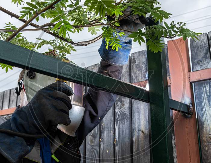 Man Welder In Welding Mask, Building Uniform And  Protective Gloves Brews Metal Welding Machine On Street Construction, In The Background Fence