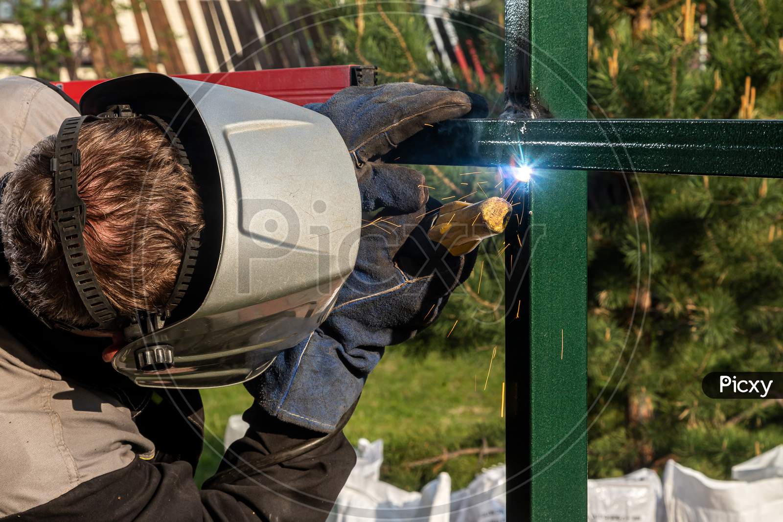 Close Up Of A Young  Man Welder In  Uniform, Welding Mask And Welders Leathers, Weld  Metal  With A  Welding Machine At The Construction  Of A Fence In Summer Day Putside In The Village