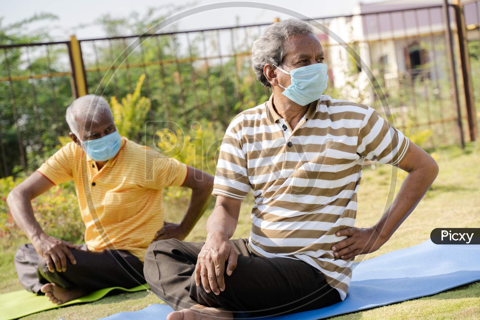 Two Active Senior People With Medical Face Mask Exercising On Yoga Mat During Early Morning - Concept Of New Normal, Elderly Helthcare And Fitness Class At Park During Coronavirus Covid-19 Pandemic