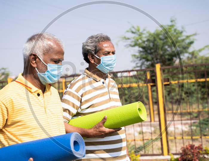 Two Senior Men With Medical Face Mask Holding Yoga Mat Coming To Park For Exercising During Morning - Concept Of New Normal Healthcare And Fitness During Coronavirus Covid-19 Pandemic.