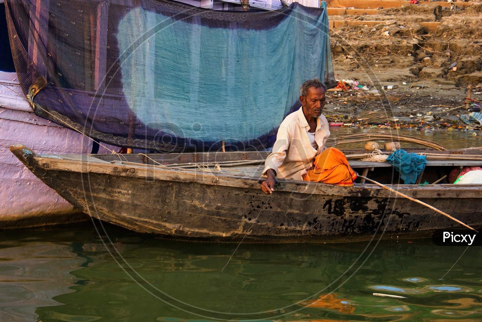 Varanasi, India - November 01, 2016: An Old Man Sitting Alone On A Wooden Boat Fishing For Living Against A Cloth Being Dried In The City Of Banaras Located In Uttar Pradesh State.