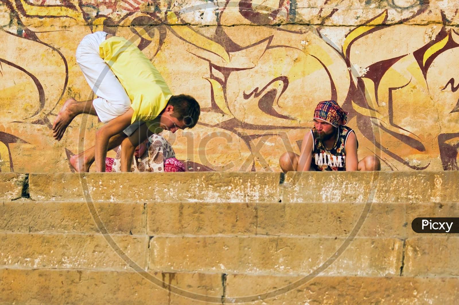 Varanasi, India - November 01, 2016: Couple Of Foreigner Men In Hipster Style Doing Yoga In The Morning With A Trainer Against Wall With Graffiti In The City Of Banaras Situated In Uttar Pradesh