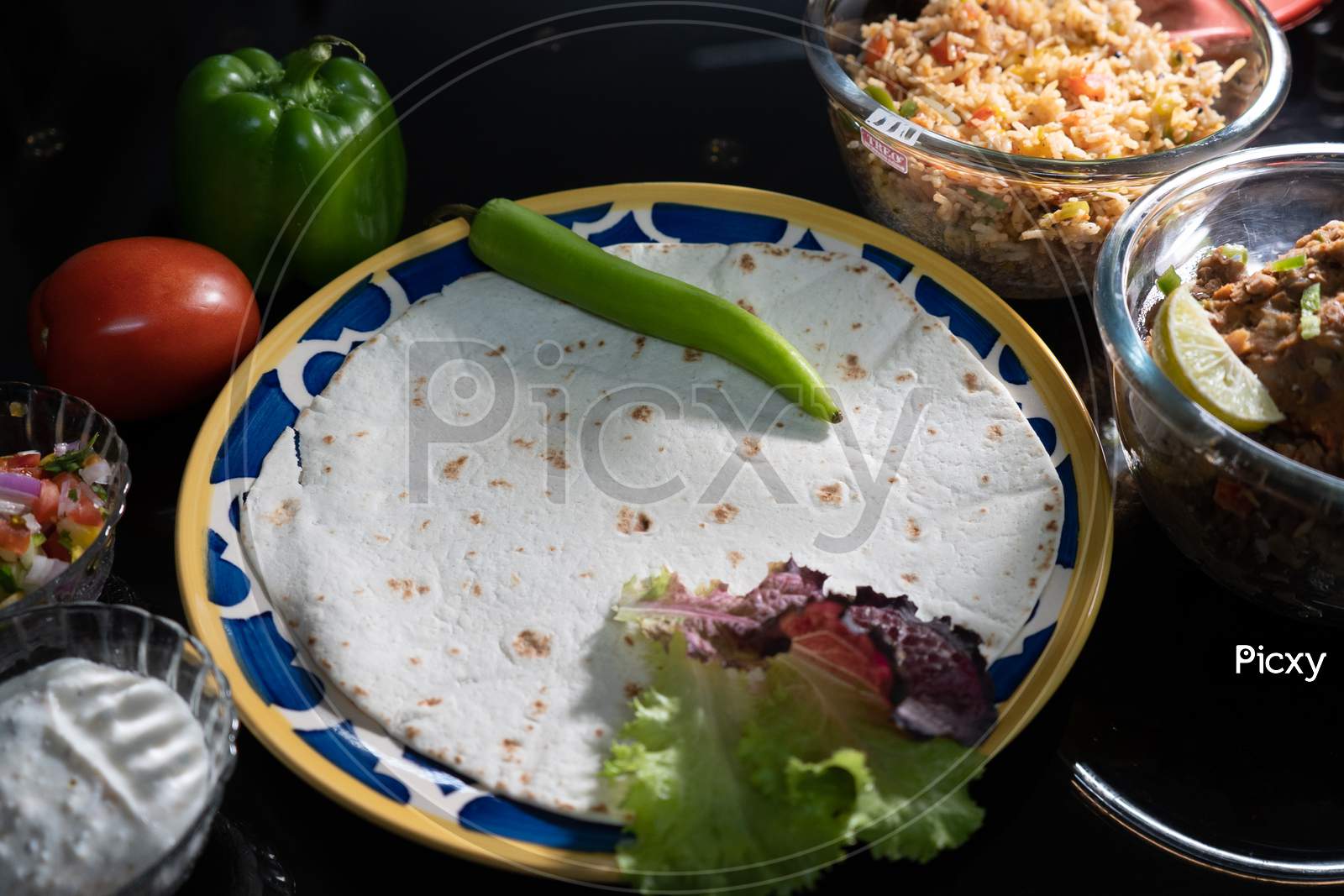 Low Key Moody Shot Showing Roti Paratha Tortilla Flatbread On Glass Plate Surrounded By Fresh Vegetables Like Capsicum, Tomatos, Rice And Meat All The Ingridients To Prepare Mexican Food