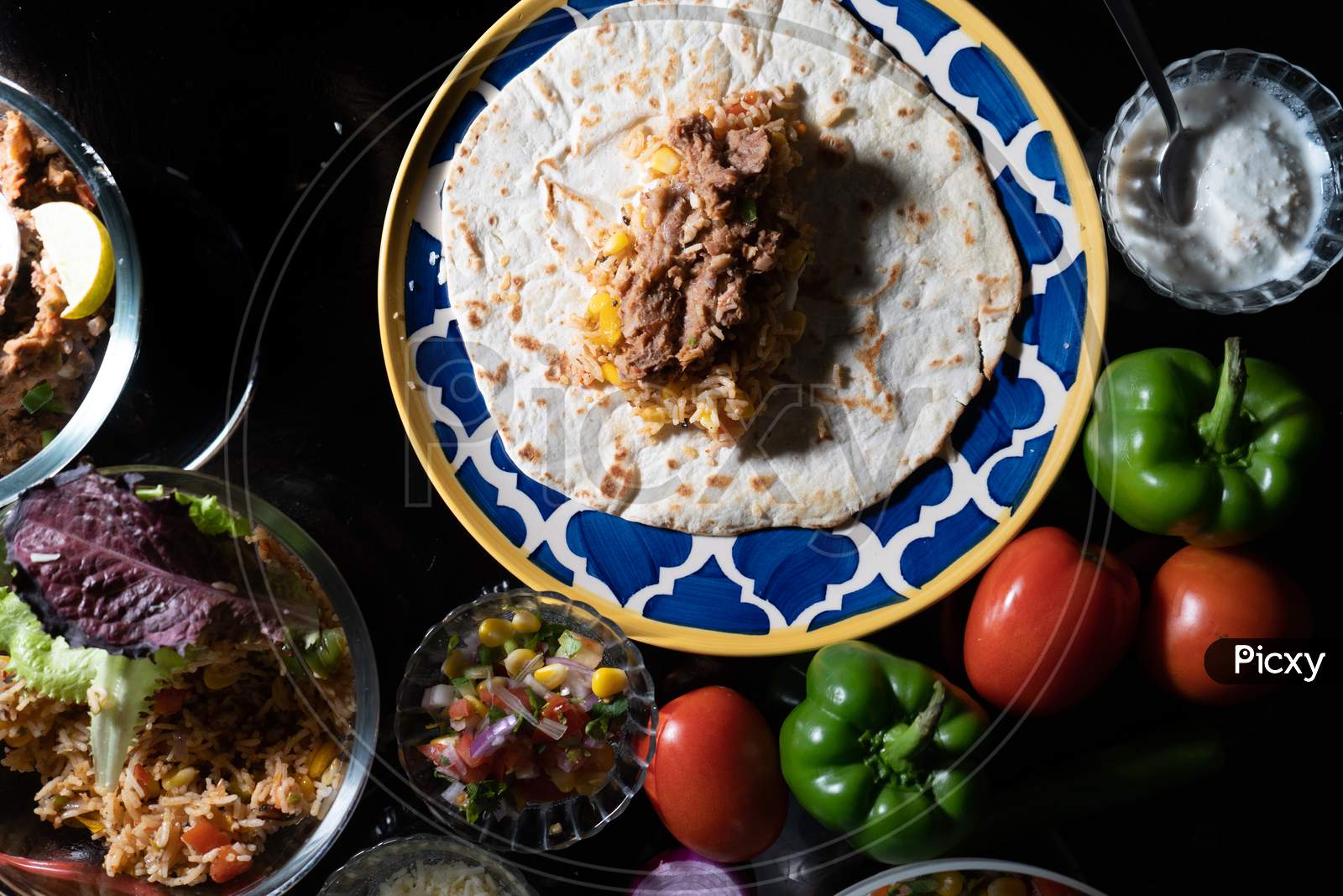 Top Flatlay Low Key Moody Shot Showing Roti Paratha Tortilla Flatbread On Glass Plate Surrounded By Fresh Vegetables Like Capsicum, Tomatos, Rice And Meat All The Ingridients To Prepare Mexican Food