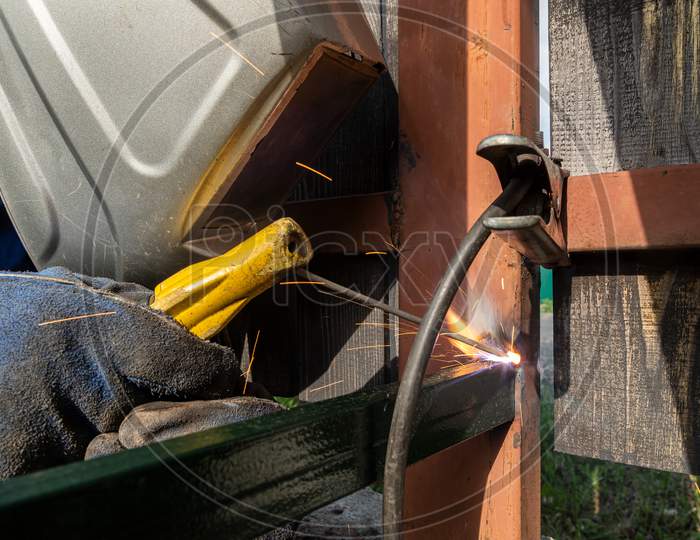 A Strong Man Is A Welder In A Welding Mask And Welders Leathers, A Metal Product Is Welded With A Welding Machine In The Outside, Yellow Sparks Fly To The Sides